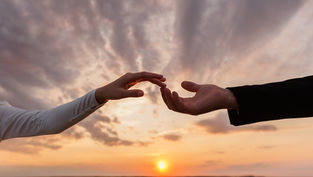 Two hands reaching toward each other at sunset, set against a dramatic sky. One hand is in a dark sleeve, the other in white.