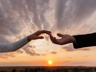 Two hands reaching toward each other at sunset, set against a dramatic sky. One hand is in a dark sleeve, the other in white.
