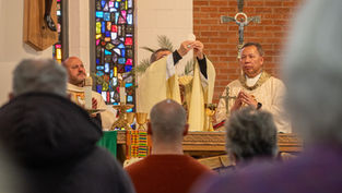 Priest elevates the host during a church service. A colorful stained glass window and attendees in the foreground emphasize reverence.