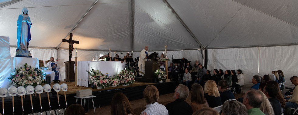 People sit and stand inside a large tent during a liturgical celebration with flowers, candles and a Marian statue near the altar.