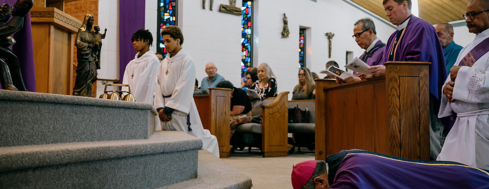 A religious ceremony with a bishop prostrate on the floor, two altar servers in white, and clergy in purple robes. Stained glass and statues in church.