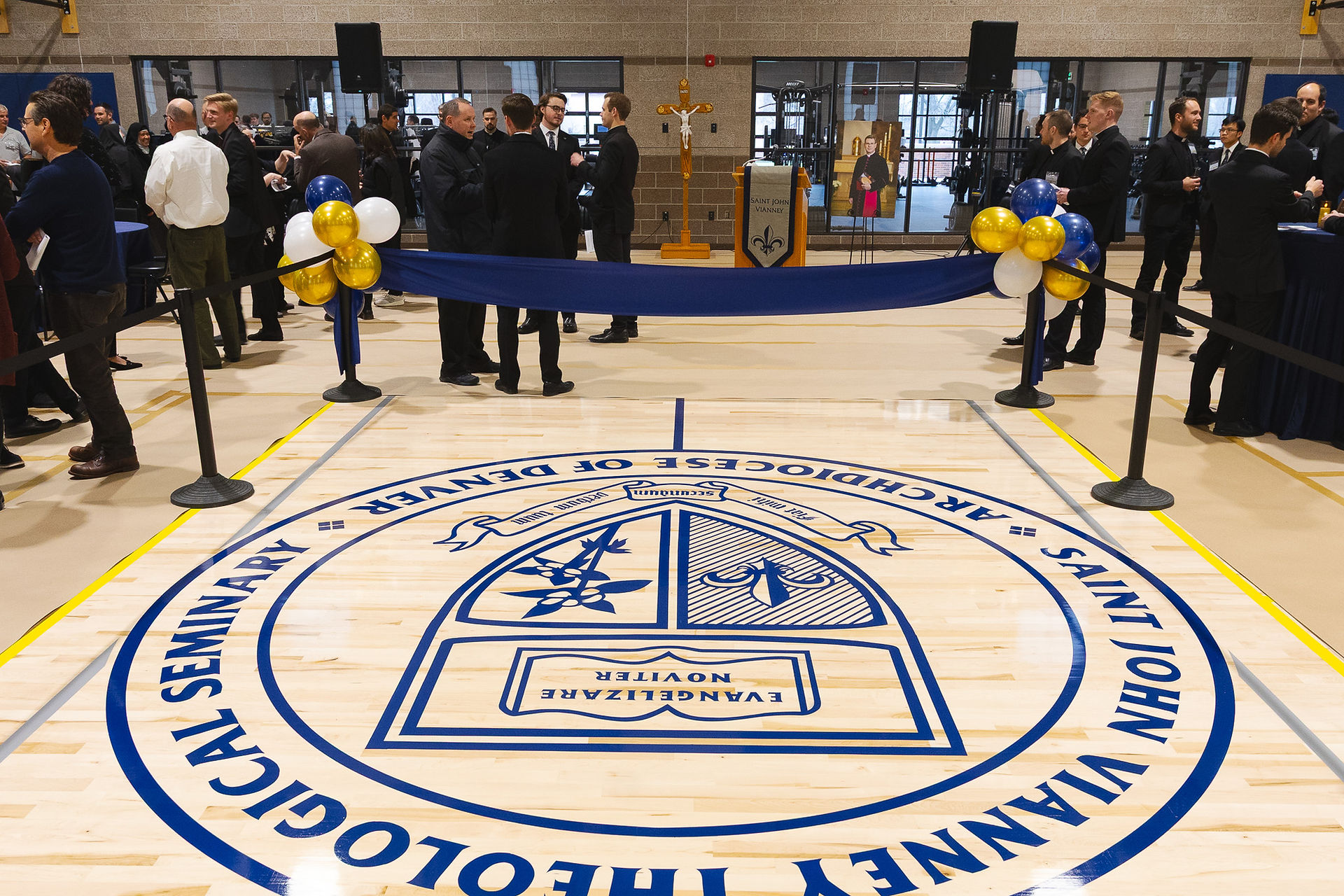 People in formal attire gather in a gym with a large "Saint John Vianney Theological Seminary" emblem on the floor. Blue and gold balloons adorn a ribbon.
