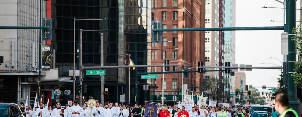 Procesión religiosa con sacerdotes de blanco y fieles en una avenida urbana, bajo letreros de 16th Street y Cleveland.