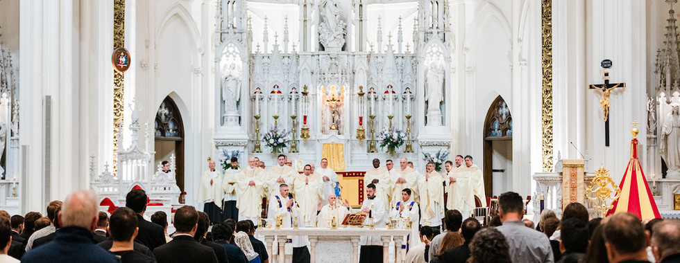 A congregation in a grand church watches priests in white robes at an ornate altar. The setting is serene with high ceilings and statues.