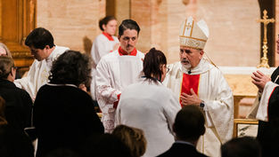 Varios clérigos en vestimentas ceremoniales realizan una bendición en una iglesia. Un altar con una cruz dorada está al fondo. Ambiente solemne.