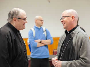 Two Catholic priests smile and talk together in a meeting room, with another man standing in the background.