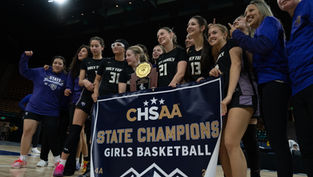 Girls' basketball team celebrates championship win, holding trophy and "State Champions" banner in stadium. Team wears black uniforms.