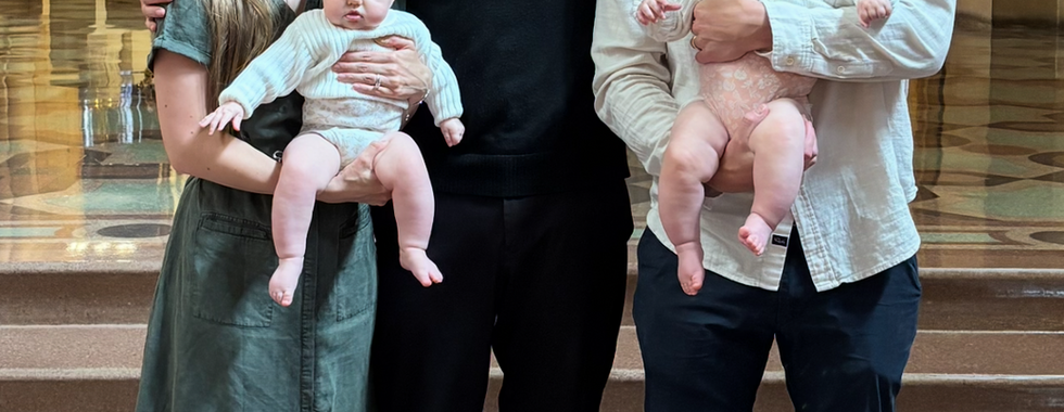 A couple holding babies with bows stands beside a priest inside a church with ornate golden decor and stained glass windows, all smiling.