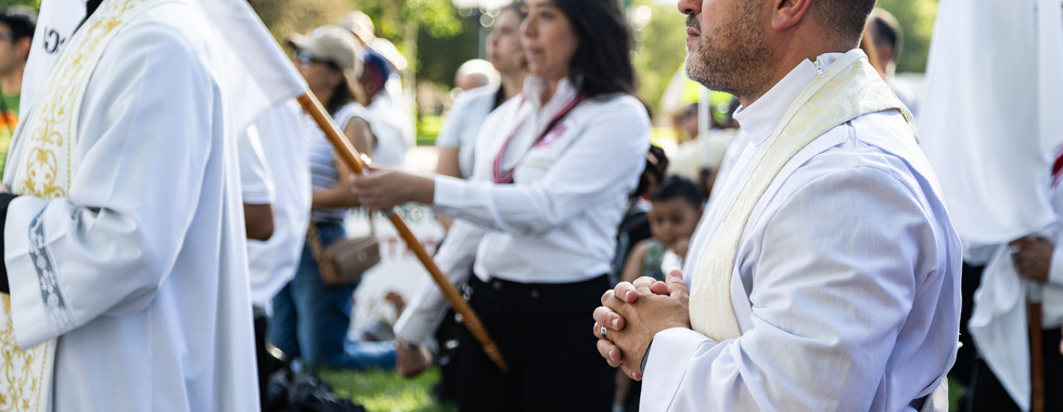 Procesión religiosa al aire libre: un hombre con túnica blanca reza con las manos juntas, rodeado de fieles y banderas.