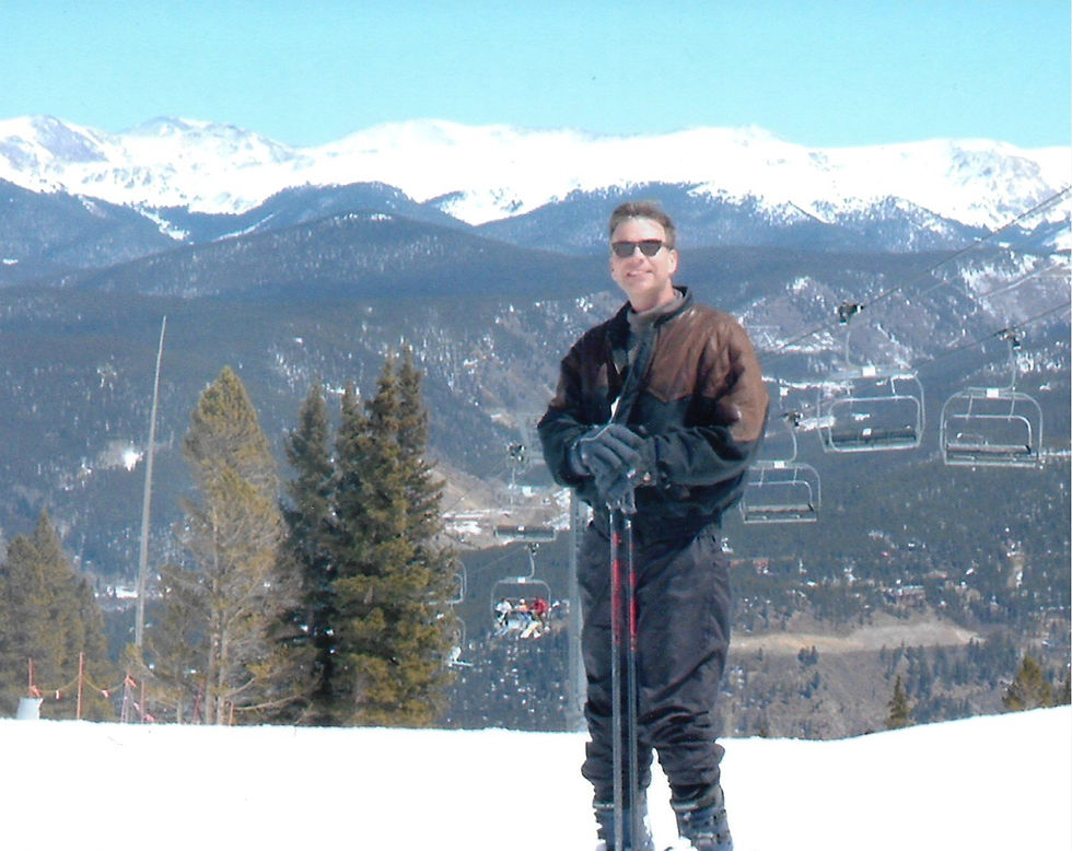Smiling person in ski gear holding poles on a snowy mountain with evergreen trees, ski lifts, and snowy peaks in the background. Sunny day.