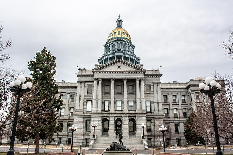 State Capitol building with a gold dome, gray stone facade, and statues. Overcast sky with trees and lampposts surrounding the entrance.