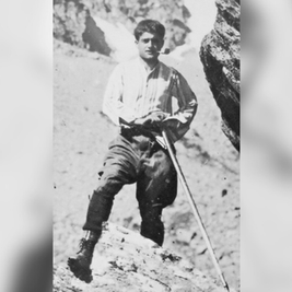 Man in period clothing leans on a walking stick in rocky terrain. He looks determined, set against a rugged mountain backdrop. Black and white photo.