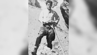 Man in period clothing leans on a walking stick in rocky terrain. He looks determined, set against a rugged mountain backdrop. Black and white photo.