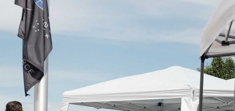 Man in uniform speaks at podium under white canopy. Audience seated outdoors. Black flag, blue sky, and person in logo shirt visible.