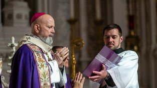 Un sacerdote con mitra y atuendo púrpura con bordados reza, mientras un hombre sostiene un libro morado. Escenario religioso solemne.