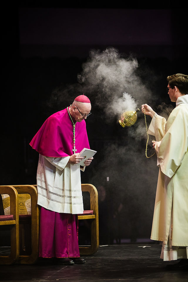 A bishop in a magenta cassock bows, holding a book. A priest swings an incense burner, smoke rising, in a solemn religious ceremony.