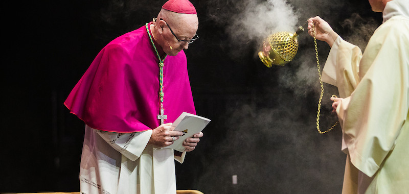 A bishop in a magenta cassock bows, holding a book. A priest swings an incense burner, smoke rising, in a solemn religious ceremony.