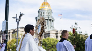 Catholic procession with priests carrying a gold monstrance and red candle before a domed capitol building under a bright sky.