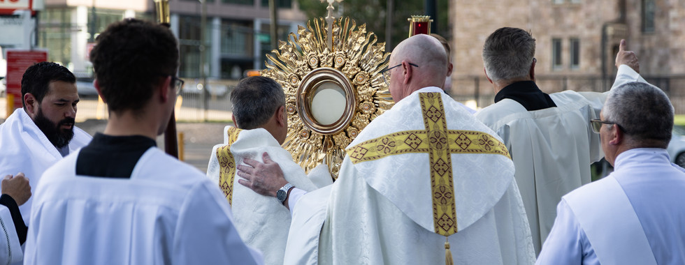 Catholic priests in white vestments process behind a gold monstrance on a city street, a solemn outdoor ritual