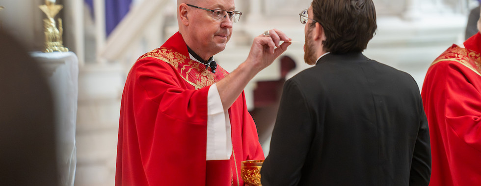 Clergyman in red robes offers communion to a man in a church. Ornate gold chalice in hand, solemn mood, blurred background.