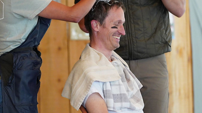 Woman applies hair dye to seated man, smiling. Man with beard speaks into mic. Indoors, wooden backdrop, casual and jovial scene.