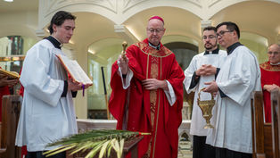A religious ceremony with clergy in red and white robes; a man sprinkles holy water over palm fronds. Elaborate arches in the background.