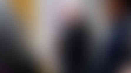 Bald man in black suit prays with a microphone, near banner with "Jesucristo". Floral arrangement and water bottles visible. Indoor setting.