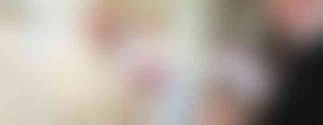 A bishop in ornate robes with a gold staff smiles at a child held by a woman in a church. Clergy and congregation are visible in the background.