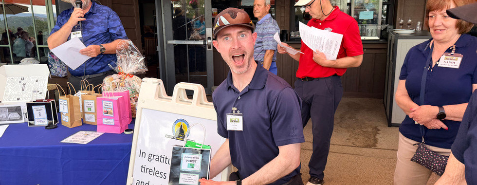Smiling man kneels holding a prize box at a church fundraiser table outside a restaurant, with volunteers and an Order Here sign behind.