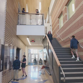 Students in a school hallway, some standing, some walking. Brick walls, staircase, and informational panels create a modern academic setting.