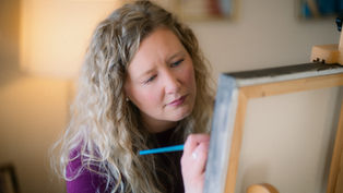 Woman with curly hair painting on a canvas in a warmly lit room, focused expression, blue brush in hand, wearing a purple top.