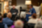 Two men sit in armchairs having a conversation. One holds a book in a warmly lit room. An audience listens attentively in the foreground.