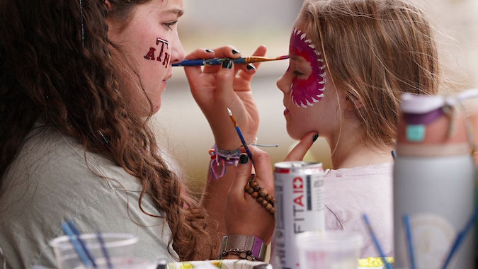 Girl paints another girl's face at a table with blue checkered cloth. The painted girl has vibrant pink face art. Fun, artistic setting.