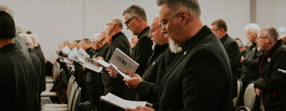 Men in black attire stand in a meeting room holding papers, likely in a solemn gathering. Rows of chairs and soft lighting in background.