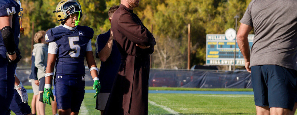 Football players and a man in a brown robe on the sideline; blue sky, trees in the background, scoreboard visible, conveying focus.