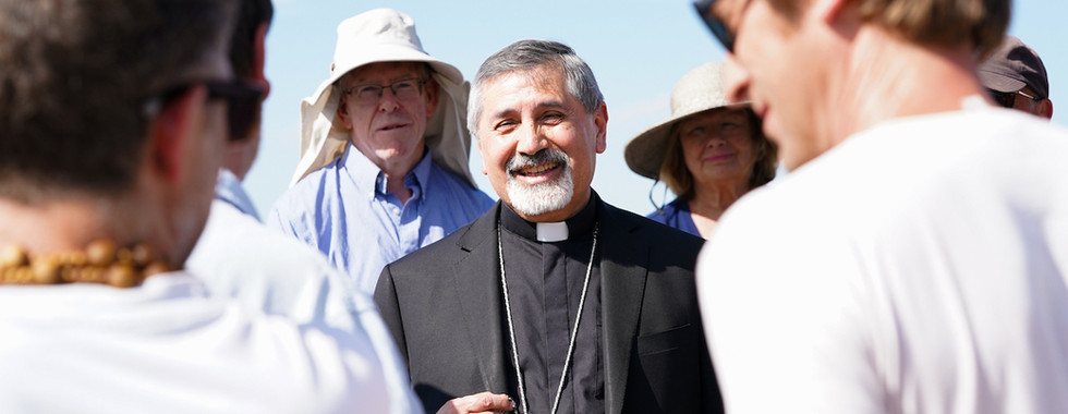 Smiling priest in black clerical clothes with a cross necklace talks with a small group outdoors under a bright blue sky.