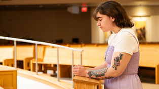 Young person in a lavender dress and tattoos prays with eyes closed in a quiet, wooden pew-filled room, exuding tranquility.