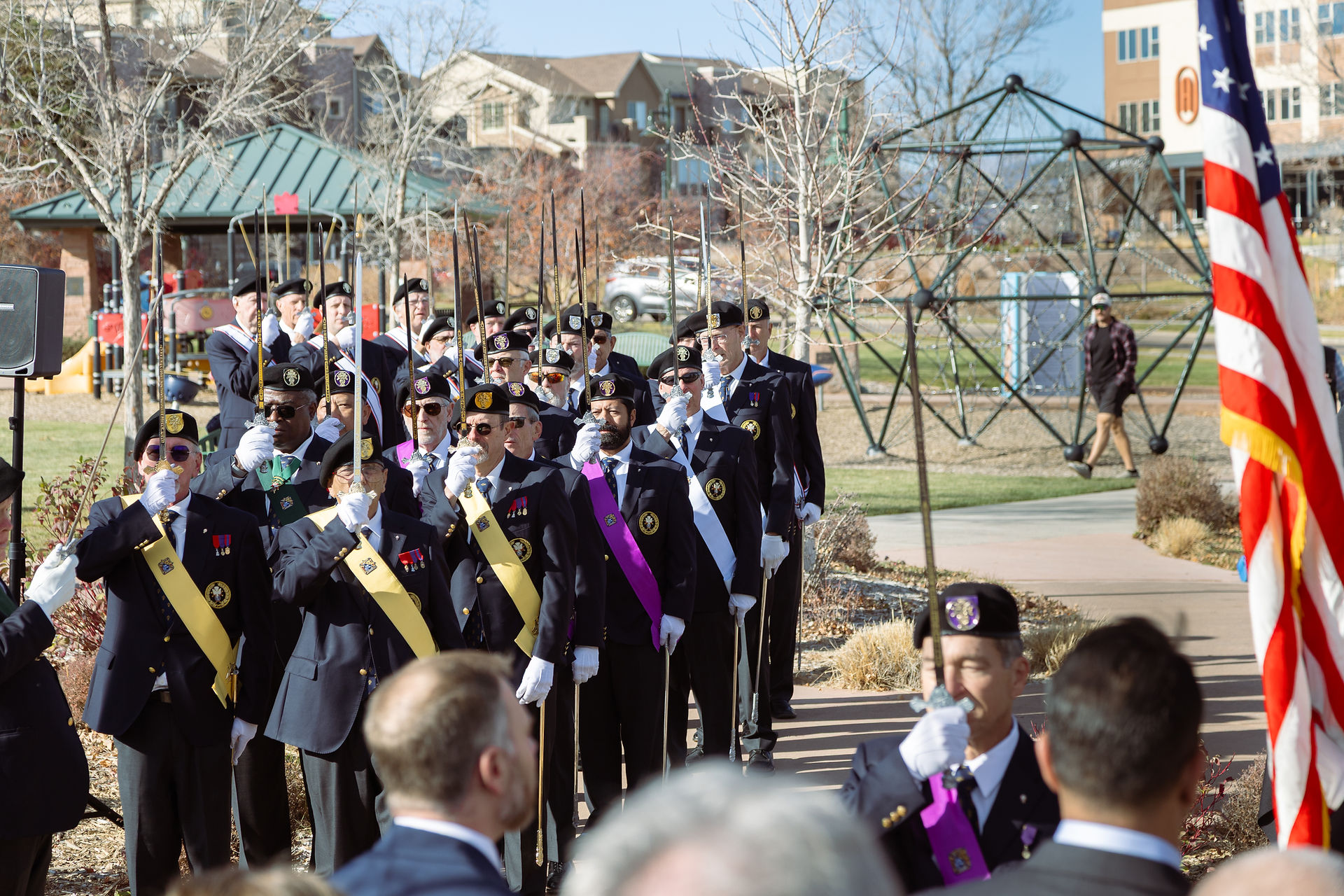 Uniformed men in a ceremony march with swords raised, wearing sashes, outdoors near a playground. An American flag is visible.