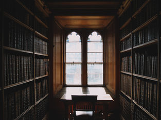 Dim library with tall bookshelves, a wooden desk, and arched window light, creating a quiet, moody atmosphere.