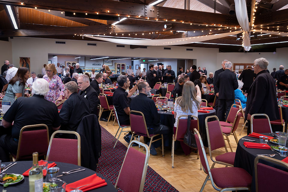 People in formal attire sit at round tables with red chairs and napkins, attending an indoor banquet under string lights, creating a warm atmosphere.