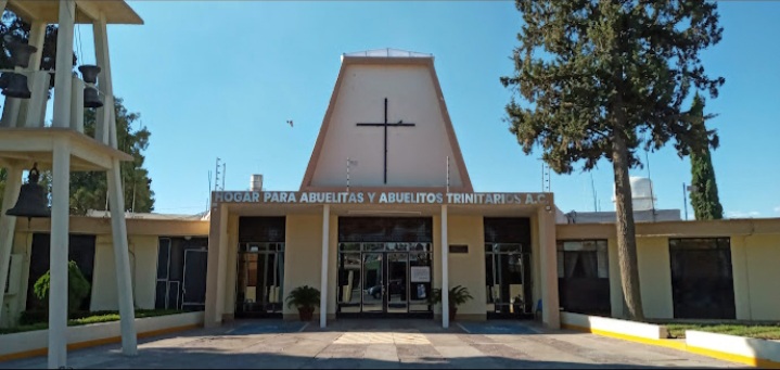 Building facade with a cross on top, sign reading "Hogar para Abuelitas y Abuelitos Trinitarios A.C." Sunny day, trees and bells visible.