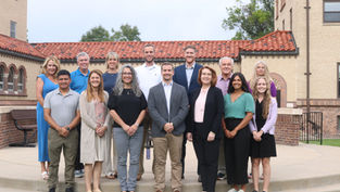 Twelve smiling adults pose on steps outside a brick building with a red tile roof, dressed in business casual.