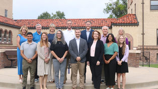 Twelve smiling adults pose on steps outside a brick building with a red tile roof, dressed in business casual.