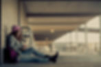 Girl in a pink beanie reads a book, sitting against a wall in a corridor. Her backpack rests beside her. The scene is calm and serene.