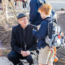 A man in clerical attire talks to a boy with a backpack outdoors. The setting is around leafless shrubs, with a sunny, cheerful mood.