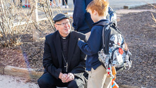 A man in clerical attire talks to a boy with a backpack outdoors. The setting is around leafless shrubs, with a sunny, cheerful mood.