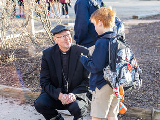 A man in clerical attire talks to a boy with a backpack outdoors. The setting is around leafless shrubs, with a sunny, cheerful mood.