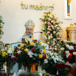 Bishop speaking at a podium surrounded by colorful flowers. Text "tu madre" on the wall. Church setting with ornate decor.