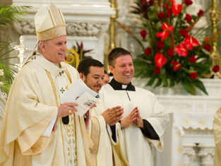 Sacerdotes en vestimentas blancas celebran en una iglesia decorada con flores rojas. Ambiente solemne y alegre.