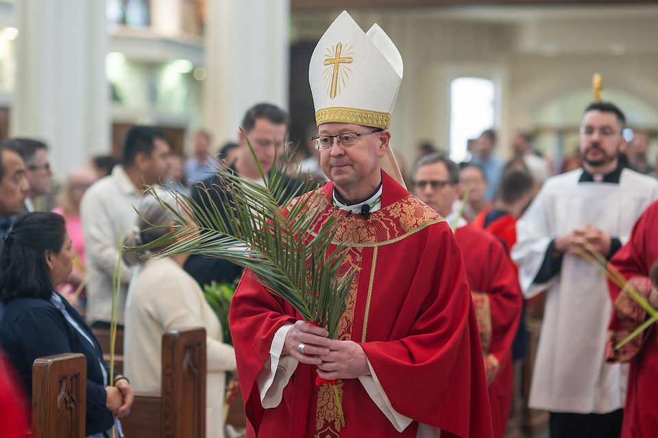 A bishop in red robes holds palm leaves during a church procession. A crowd in pews, subdued lighting, and ceremonial mood are visible.